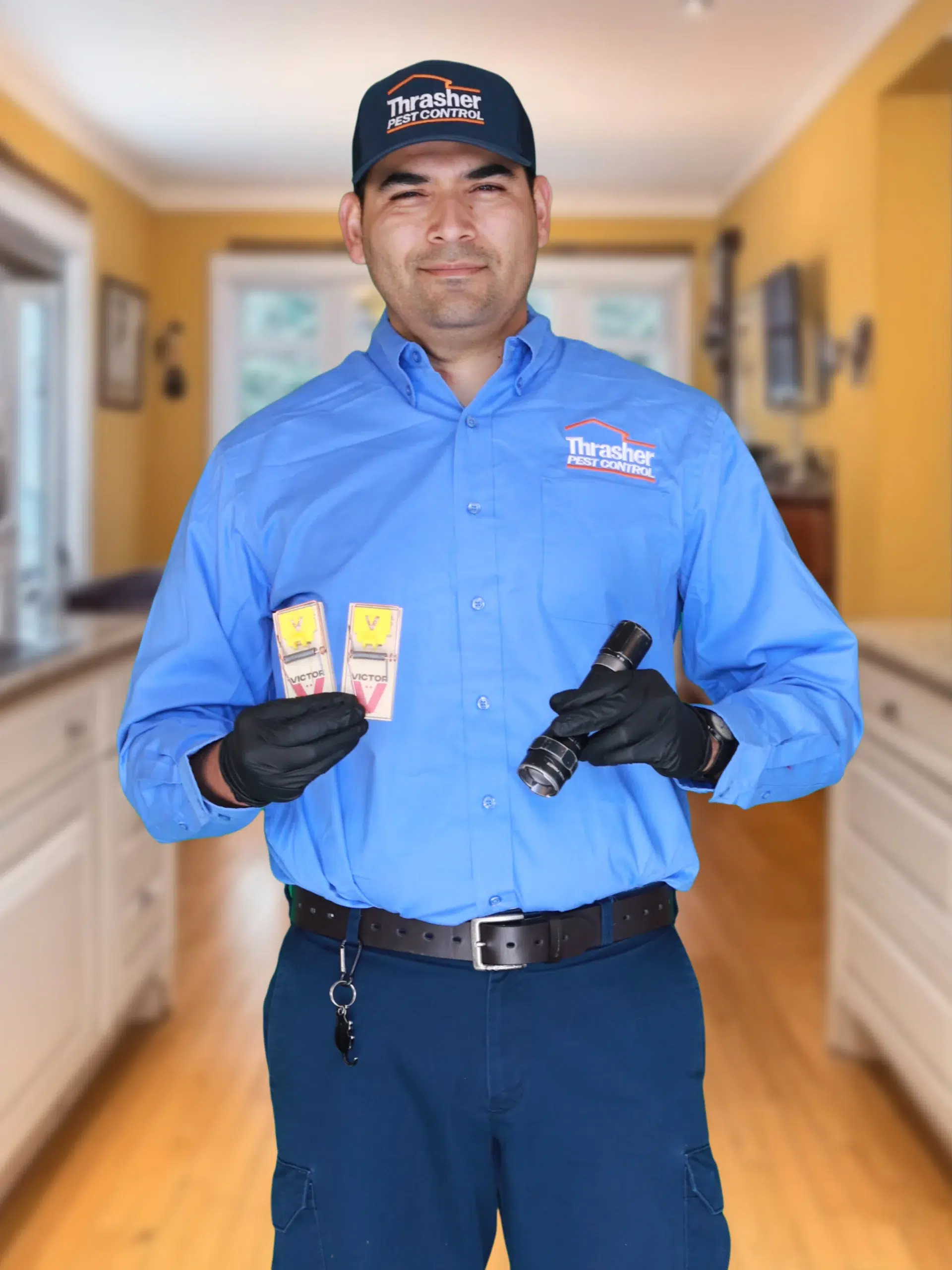 Technician in Thrasher Pest Control shirt and cap holding two mouse snap traps and a flashlight in a kitchen